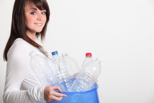 A woman with shoulder-length brown hair and a light complexion is smiling slightly while holding a blue plastic container filled with several empty, transparent plastic bottles, some with blue and red caps. She is wearing a long-sleeved white top and stands against a plain white background. The bottles in the container vary in size and shape and appear to be used for soft drinks or water. The image is related to waste collection or rubbish removal services, illustrating the collection of recyclable plastic waste. The scene is well-lit with even lighting, emphasizing the clear details of the bottles and the woman's expression. The background offers no additional environmental context, focusing solely on the woman and the container of bottles, which may be relevant to services offered by Garden Clearance Mayfair, operating in the Mayfair area, for efficient rubbish removal and recycling in London.