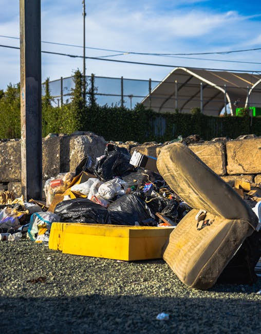 A man with dark hair, wearing a black T-shirt with white text and graphics, is leaning over to dispose of waste into a tall, cylindrical, stainless steel rubbish bin on a paved outdoor area. He is hol