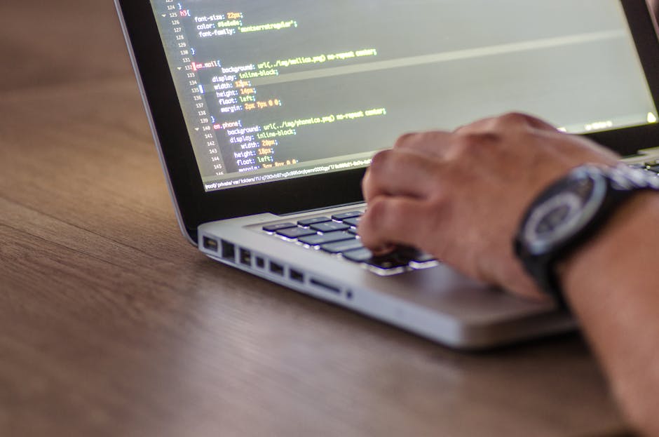 A close-up view of a person's hand typing on a silver laptop keyboard placed on a wooden surface. The screen displays lines of programming code with syntax highlighting in various colors, including ye